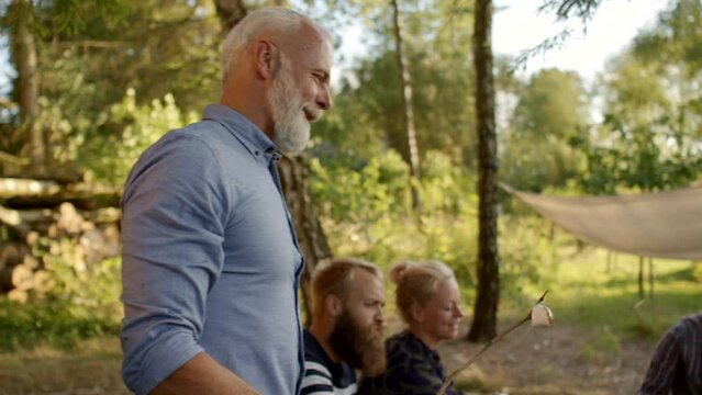 Smiling mature man standing outside with family and friends during a summer afternoon campfire
