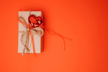 A gift wrapped in brown paper decorated with a red shiny heart on a red background. Valentine's Day. Flat lay