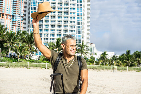 Happy male tourist in a hipster clothes waving his hat to someone on the south beach.