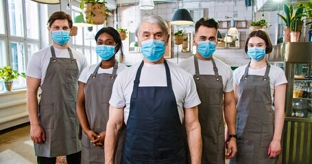 Portrait of mixed-race male and female workers in aprons and medical masks standing in cozy restaurant looking at camera in good mood waiting for customers. Business industry. Work in quarantine