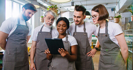 Portrait of young African American pretty female manager in apron tapping on tablet surfing internet discussing with Caucasian male and female colleagues working plan. Restaurant concept