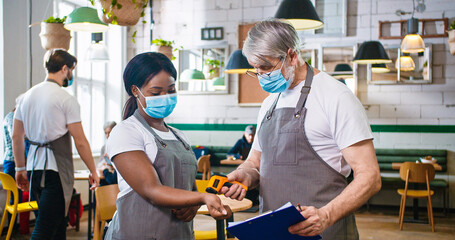 Portrait of old Caucasian man worker in cafeteria measuring temperature of young African American beautiful woman colleague in medical mask using infrared thermometer writing symptoms, health check