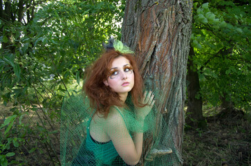 Naklejka na ściany i meble A girl in the form of a forest nymph near a tree against the background of tree bark and green foliage.