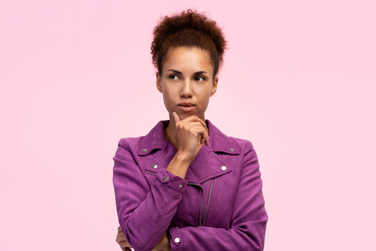 Pensive ethnic woman holding hand on her chin and thoughtfully looking aside at copy space on pink