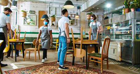 Mixed-race different people workers in coffee house working together putting chairs on tables to clean area at the end of workday. Preparing cafe before opening in quarantine. Job concept