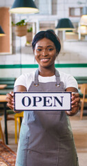 Vertical orientation of African American young beautiful female worker wearing apron standing in restaurant in apron holding in hands Open sign, waitress working in restaurant, small business concept