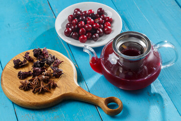 Kettle of hibiscus tea with cherries next to the table on a wooden board anise and cinnamon and hibiscus flowers and cherries in a plate. © finist_4