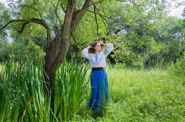 A girl in a long blue skirt and an embroidered shirt among the green meadow and marsh grass and willow trees.