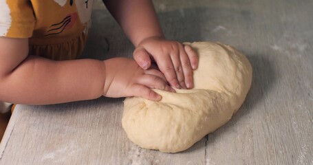 A little girl in a dress with her hands kneads the dough at home on the kitchen table. The family is doing what they love. Mom and daughter spend time preparing dough.