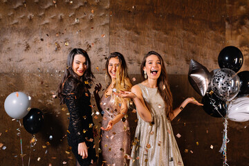 Excited lovely cute girls in festive dresses dancing and having fun under confetti with balloons on dark background in studio. happy girls celebrating, spend time together