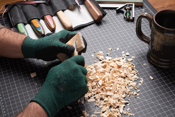 Carving tools, knife removing wood shavings from a block of basswood. Cutting mat with shavings, gloves, knives, and coffee mug in background.