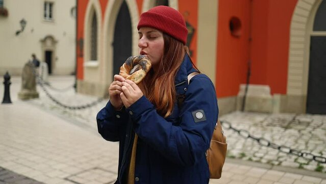 Fall tourist woman in a bright hat and autumn jacket holding and eating baked obwarzanek traditional polish cuisine snack bagel on old city Market square in Krakow. High quality FullHD footage