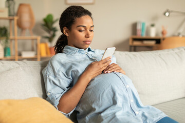 African american pregnant woman using smartphone, sitting on couch at home, surfing internet or...