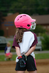 A young girl wearing a Pink Helmet with a facemask is standing on first base with her hair hanging...