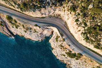 Coastal road leading along the rocky cliff and the calm sea at picturesque sunset. Aerial top down...
