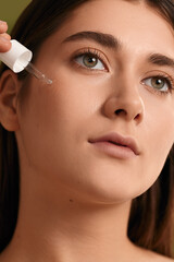 Crop young woman applying serum on face and looking away