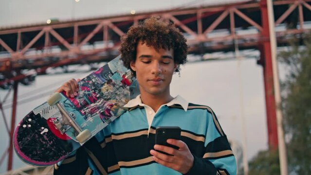 Contemporary skateboarder texting phone closeup. Curly man carrying longboard