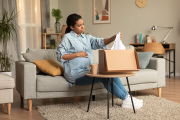 Discontented young pregnant black woman unpacking box and holding delivered clothes, sitting on...