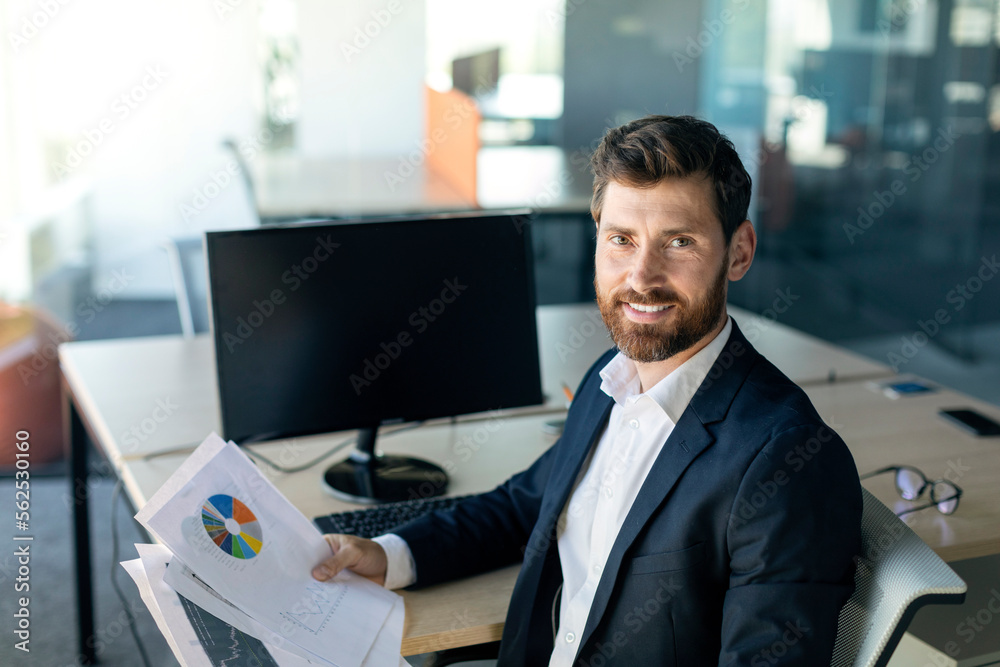 Poster portrait of positive businessman holding documents and smiling at camera, sitting at desk with blank