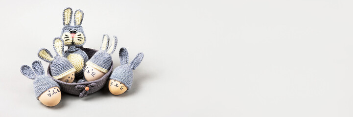 Five Easter eggs with funny faces and gray crocheted hats with bunny ears in a decorative basket and a crocheted gray bunny on a gray background. Happy Easter concept. Banner