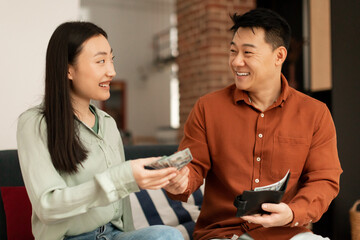 Wealthy korean mature husband giving money to his young wife, sitting together on sofa at home