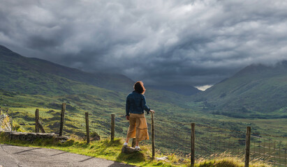 Fototapeta na wymiar Young woman in a dress enjoys the unforgettable views of Killarney National Park, the dramatic skies and green mountains of the Ring of Kerry near the town of Killarney in County Kerry, 