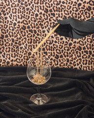 Black gloved hand holding chopsticks, picking gold necklace from wine glass against black and cheetah print background. Angled composition.