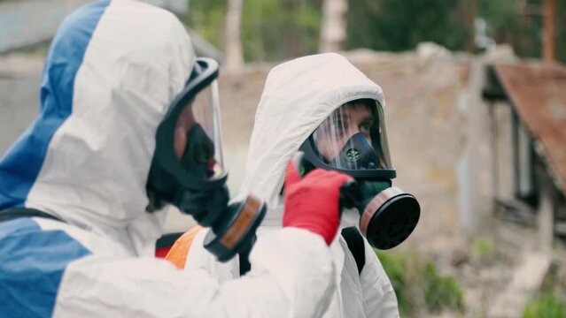 Skilled disinfection workers wearing full face respirators and white protective suits talk near abandoned old buildings closeup