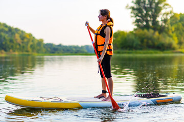 woman in life jacket at sub board at river ar evening , forest trees background