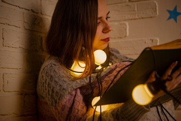 Young woman wrapped in garland of glowing bulbs reading book, sitting on floor in room. Female sitting in dark with light on bulbs turned on, relaxing with book.