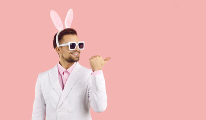Young handsome man with easter rabbit ears wearing white suit, pink shirt and sunglasses standing...