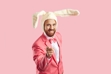 Funny young guy in Easter bunny costume points his finger at camera. Happy confident bearded man wearing plush toy rabbit ears standing on pink studio background, smiling and pointing at camera