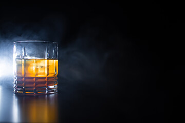glass with orange yellow liquor with ice on a black background with smoke reflection of the glass on the table with space for text