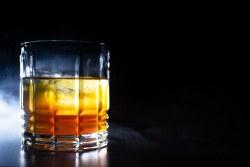 glass with orange yellow liquor with ice on a black background with smoke reflection of the glass on the table with space for text