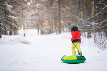 Cute happy baby runs through the winter forest and carries a snow inflatable sled. Baby in bright overalls in a snowy winter forest. Activities for children
