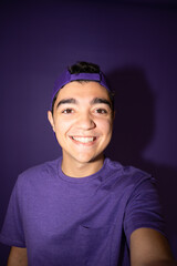 Happy hispanic teenager boy taking selfie isolated on purple background