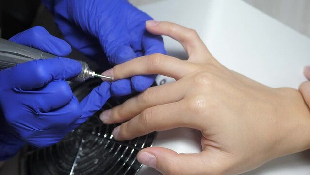 Nail master in blue rubber gloves preparating of nails for manicure. Beautician using electric nail file, cuticle pusher, to trim remove cuticles in a beauty salon. Hardware manicure milling cutter