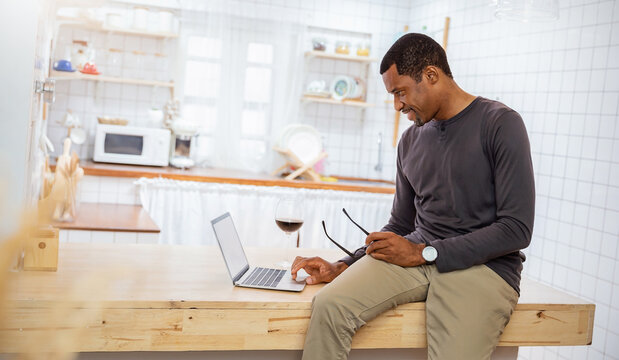 Portrait African American black man  creative relax sit on desk counter bar use computer laptop. Smile happy afro male work at table kitchen home. Freelance small business owner sme work from home.