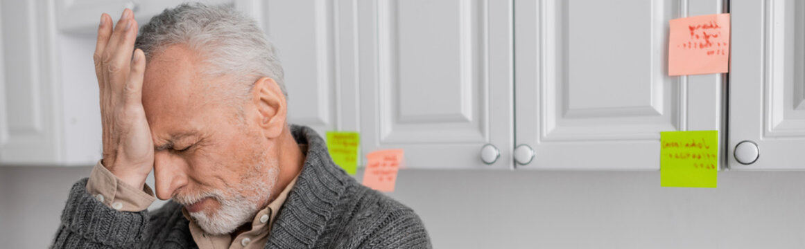 stressed man with alzheimer disease touching forehead in kitchen, banner.