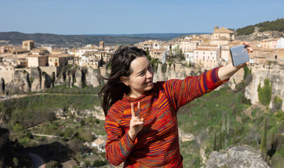 Positive interested woman recording video with mobile phone outdoor on background of Cuenca cityscape located in mountains on rocky ledge on sunny spring day