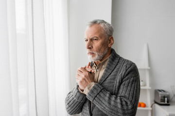 grey haired man with alzheimer disease standing with clenched hands and looking away near window at home.