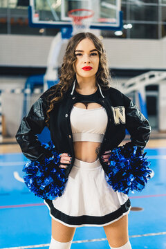 Brunette cheerleader in black and white uniform with a jacket posing with blue shiny pom-poms. Basketball court blurred in the background. High quality photo