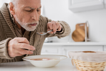 aged man with parkinson disease holding spoon in trembling hand while eating soup in kitchen.