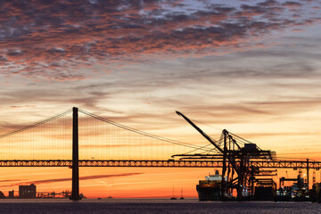 Industrial cranes, a suspension bridge, and a tourist liner sailing along the river at sunset
