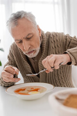 aged man with parkinson disease and hands tremor sitting with spoon near plate with soup in kitchen.