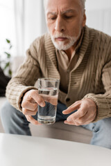 blurred man with parkinson syndrome and tremor in hands sitting and holding glass of water at home.