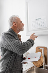 Fototapeta premium senior man with alzheimer disease holding blank notebook and pointing at calendar in kitchen.
