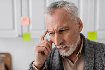 thoughtful senior man with alzheimer disease touching head near blurred sticky notes in kitchen.