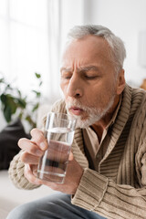 grey haired man with parkinsonism holding glass of water in trembling hands at home.