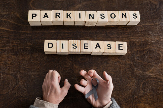top view of cropped man holding grey ribbon in trembling hands near blocks with parkinsons disease inscription on wooden surface.